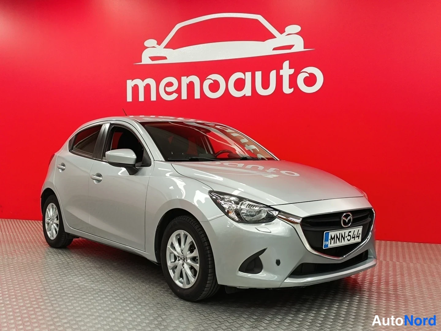 Etuviisto, nokka oikealle: Hopeanvärinen Mazda 2 -viistoperä auton etukulmasta, rekisterikilpi, punainen menoauto-tausta.