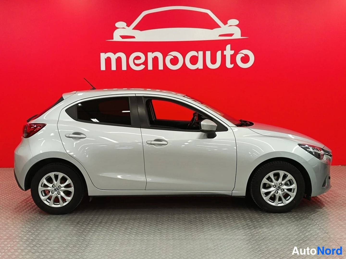 Sivulta: Hopeanvärinen Mazda 2 -viistoperä sivulta vasemmalta, punainen menoauto-tausta.