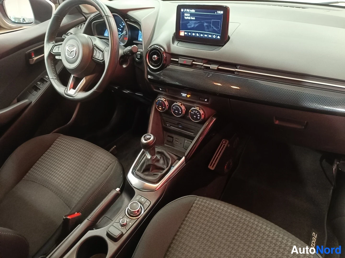Muu: Auton ohjaamo: Mazda-ohjauspyörä, viisivaihteinen manuaalivaihteisto, keskikonsolissa infotainment-näyttö ja kolme säätöpyörää.