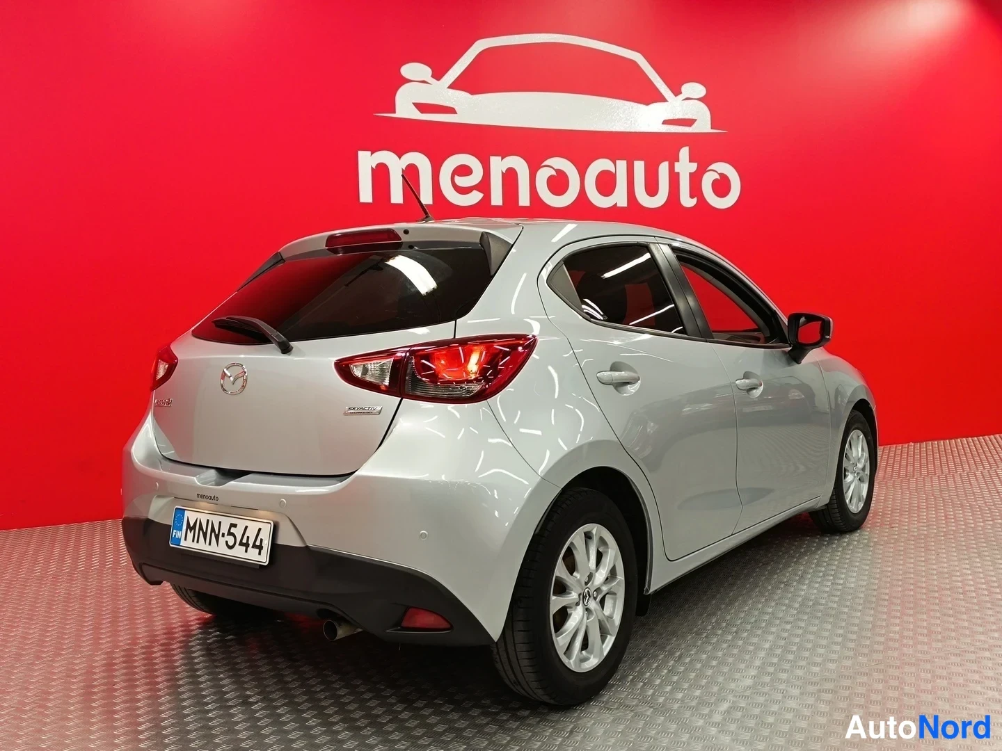 Takaviisto: Hopeanvärinen Mazda 2 -viistoperä takakulmasta, rekisterikilpi, punainen menoauto-tausta.