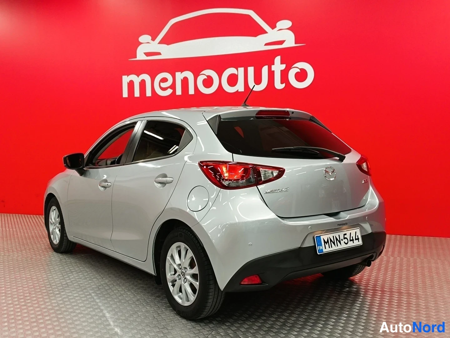 Takaviisto: Hopeanvärinen Mazda 2 takaapäin vasemmasta viistosta, rekisteritunnus, punainen taustaseinä.