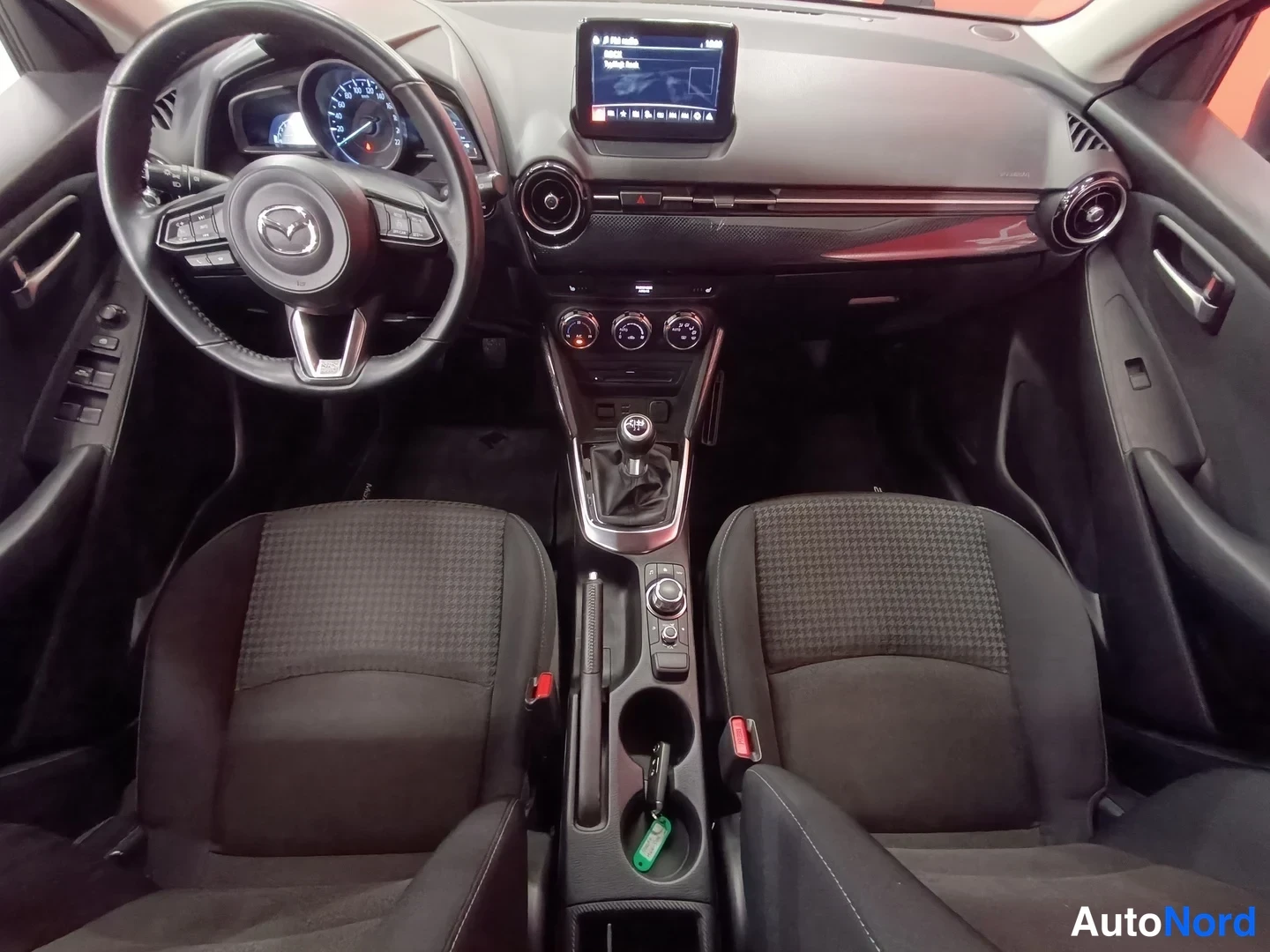 Muu: Mazda 2:n ohjaamo ylhäältä etuistuimilta: mittaristo, Mazda-logo ratti, manuaalivaihteisto ja kosketusnäyttö.
