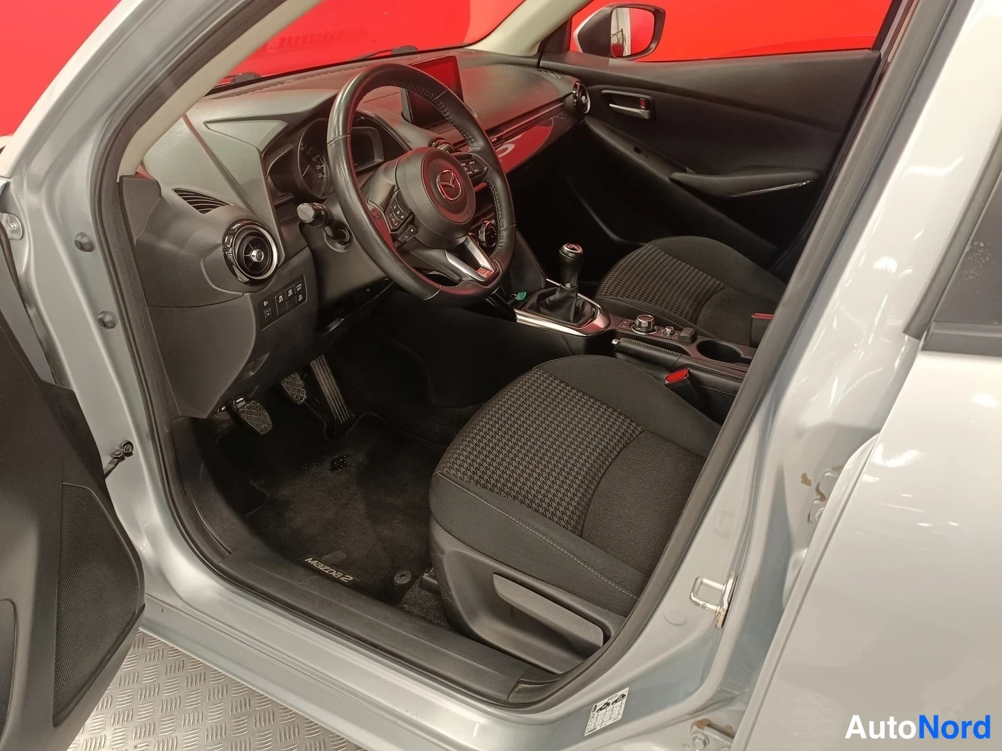 Muu: Mazda 2:n ohjaamo avoimesta kuljettajan ovesta: ratti, kytkinpoljin, manuaalivaihde ja kangasistuimet.