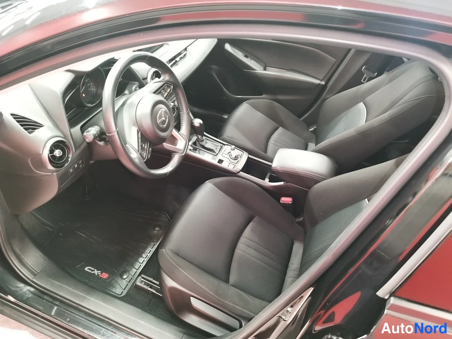 Yksityiskohta: Näkymä avoimesta etiovesta: etuistuimet, Mazda-ohjauspyörä, keskitunneli automaattivaihteella ja CX-3-merkkiset matot.