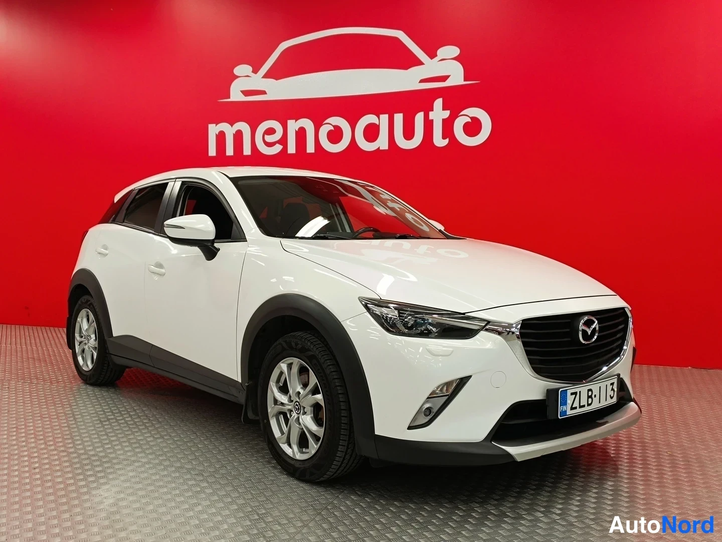 Etuviisto, nokka vasemmalle: Valkoinen Mazda CX-3 -henkilöauto kulmassa etu- ja sivunäkyvissä punaisella menoauto-taustalla.