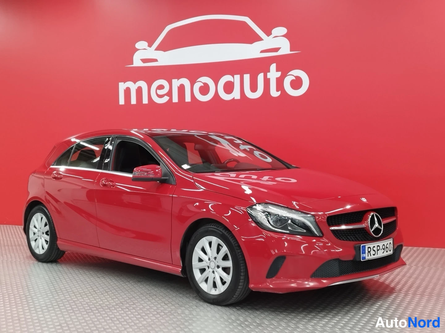 Etuviisto, nokka oikealle: Punainen Mercedes-Benz A -mallinen viistosti edestä oikealta kuvattu farmarivaline harmaalla lattialla punaisella menoauto-taustalla.
