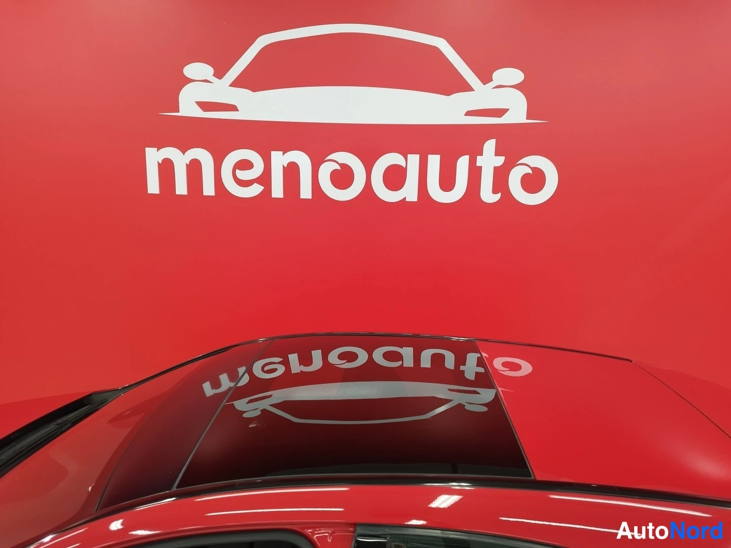 Yksityiskohta: Punaisen auton katon yläosa, kuva vino; tausta punainen menoauto-logo seinällä.