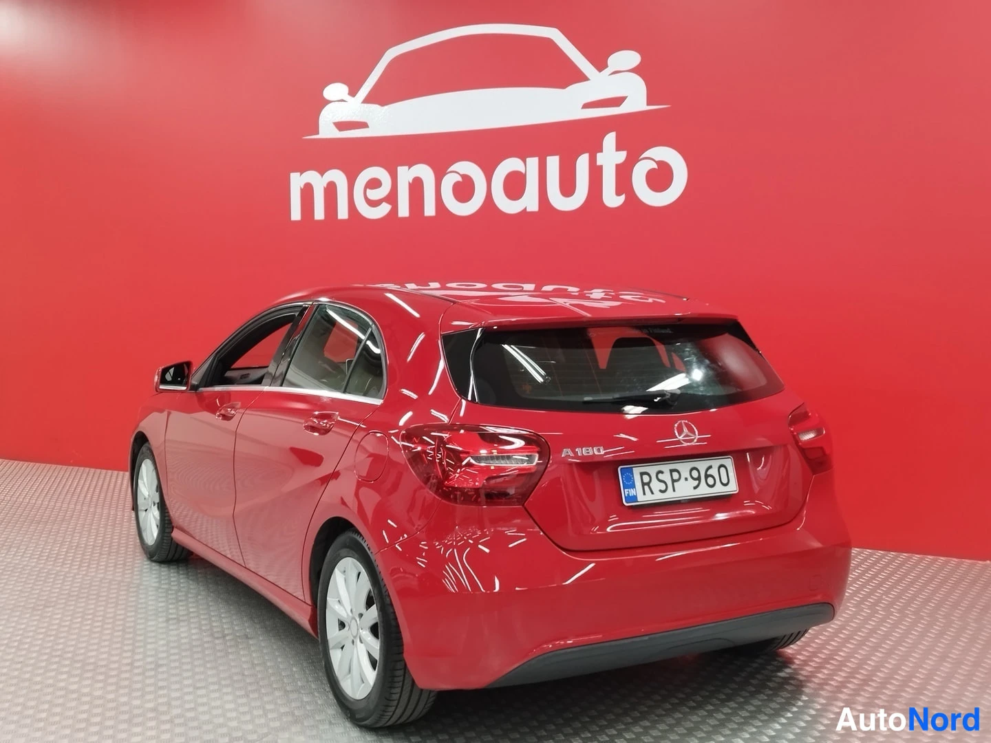 Takaviisto: Punainen Mercedes-Benz A 180 -viistoperä takaapäin kuvattuna, rekisteritunnus, autoa myyvä menoauto-taustaseinä punainen.