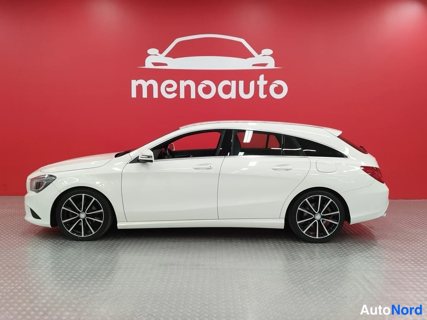 Etuviisto, nokka vasemmalle: Valkoinen Mercedes-Benz CLA Shooting Brake -farmari sivulta etukolmanneltaan, tummat kevytmetallivanteet, punainen menoauto-taustaseinä.