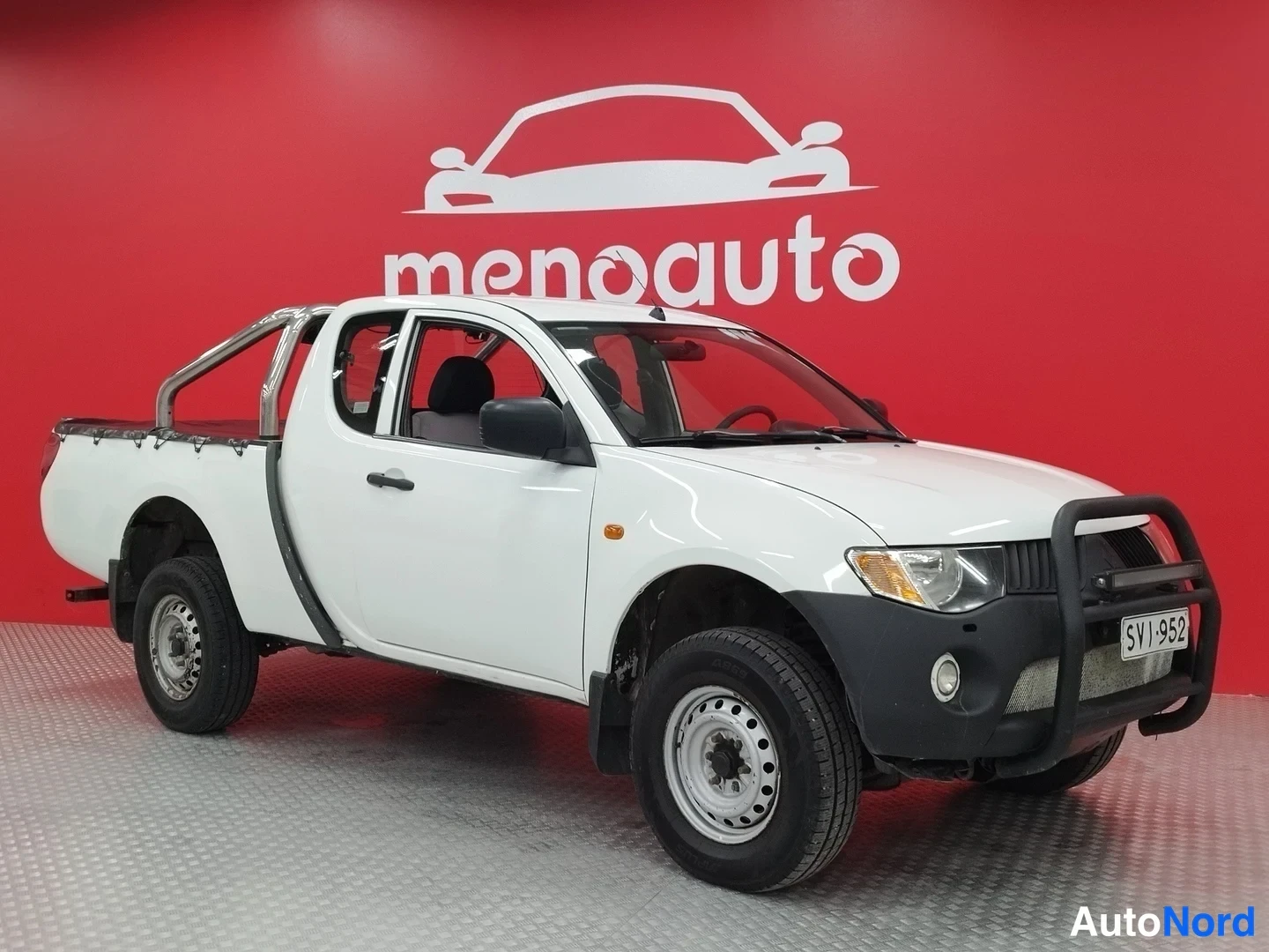 Etuviisto, nokka oikealle: Mitsubishi L200 -lainen valkoinen avolava-auto kuvattu kulmasta etuosaa ja vasenta sivua näkyen, tumma suojarauta etupuskurissa, taustalla punainen menoa