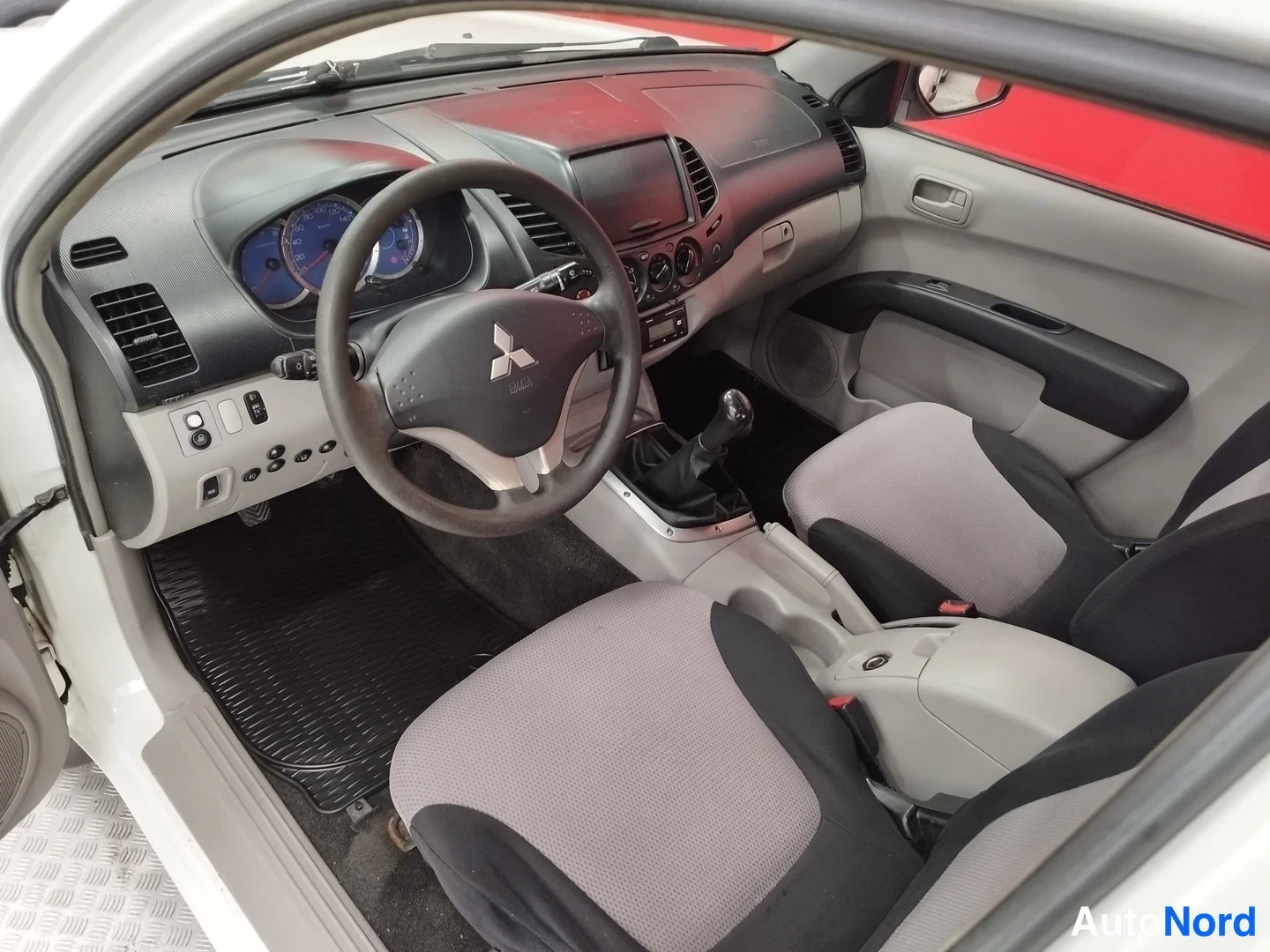 Yksityiskohta: Auton sisätilat: Mitsubishi-ohjauspyörä, analogiset mittarit, manuaalivaihteisto ja tummat istuimet.