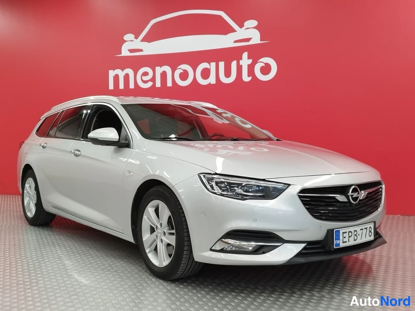 Etuviisto, nokka oikealle: Hopeanharmaa Opel Sports Tourer -asema-auto etuosa kolmannes oikealta, rekisteritunnus, punainen menoauto-taustaseinä.