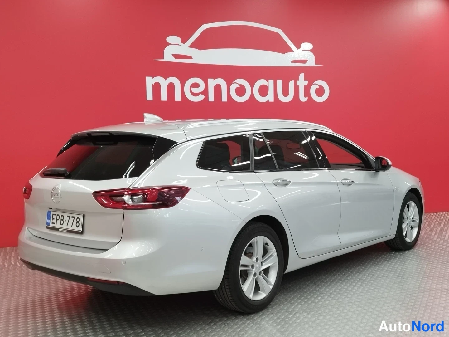 Takaviisto: Hopeanharmaa Opel Sports Tourer -asema-auto takaosa kolmannes vasemmalta, rekisteritunnus, punainen menoauto-taustaseinä.
