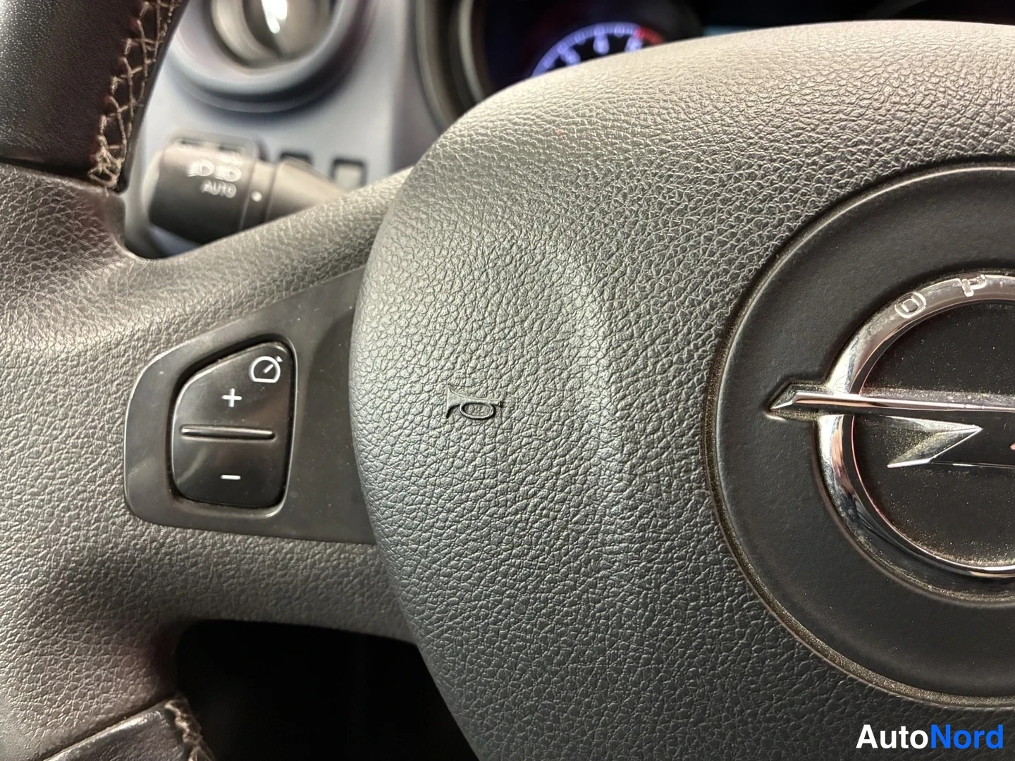 Edestä: Ohjauspyörän lähikuva Opel-merkillä ja vakiinopeudensäätimen painikkeilla (+/- ja säätökuvake).