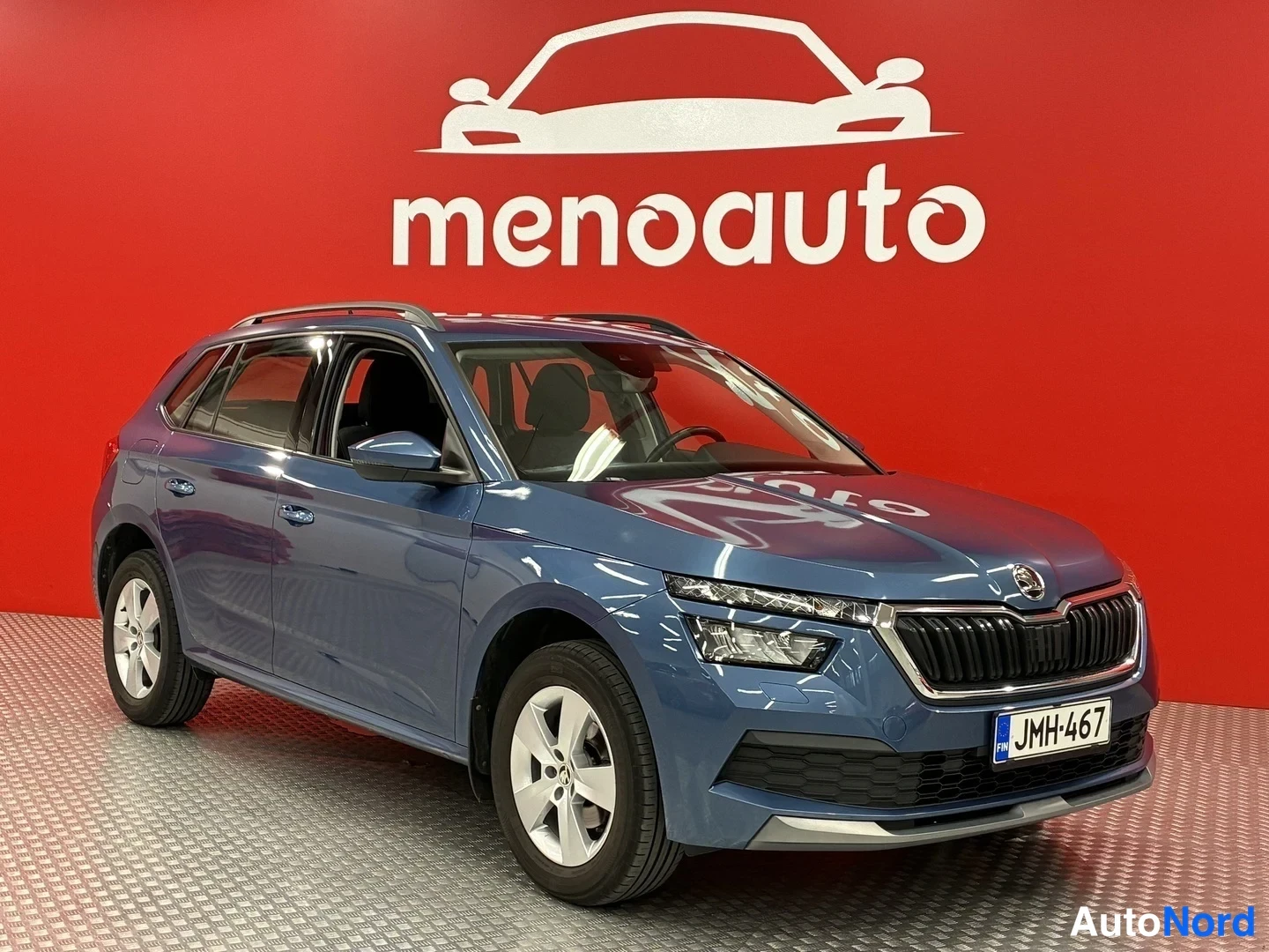 Etuviisto, nokka oikealle: Sininen Skoda Kamiq etuosa kolmesti oikealta kuvattuna punaisella menoauto-taustalla, rekisteritunnus.