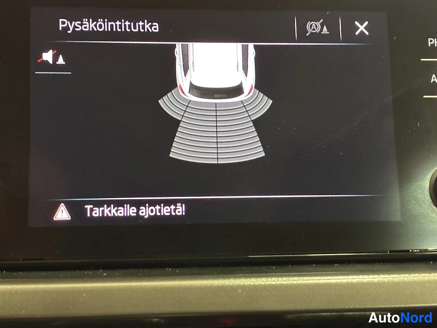 Yksityiskohta: Infonäytön pysäköintitutkan näyttö: valkoinen auto kuvasta ja teksti 'Tarkkaile ajotietä!'