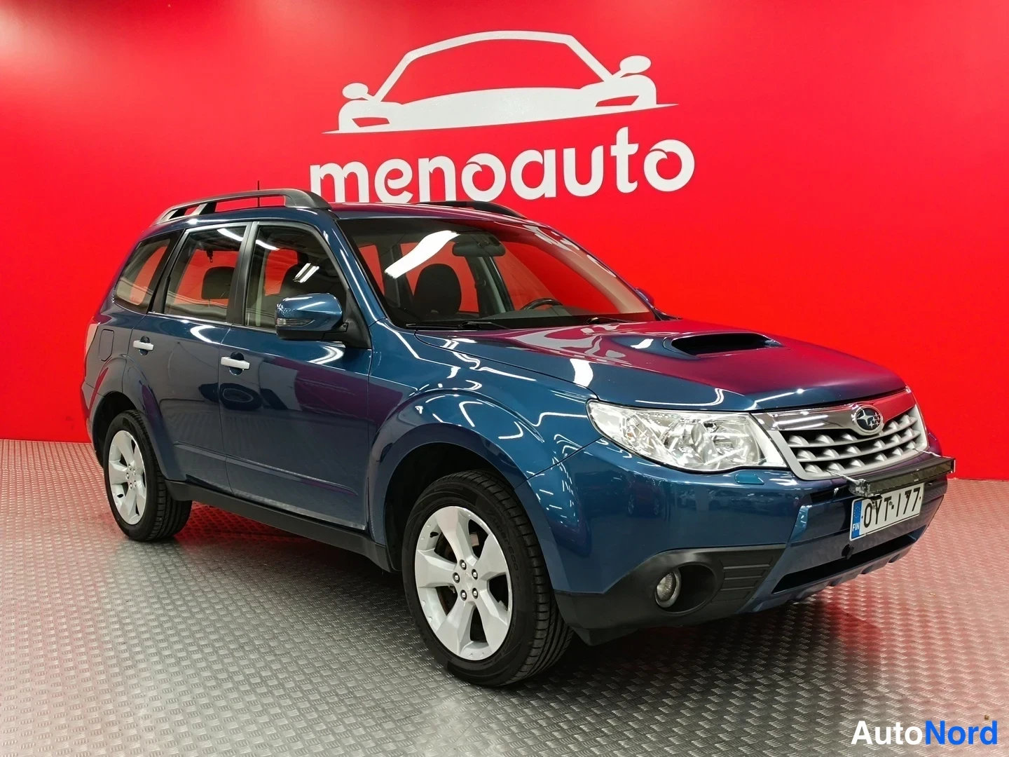 Etuviisto, nokka oikealle: Sininen Subaru Forester esillä punaisella menoauto-taustalla, etukulmasta oikealta
