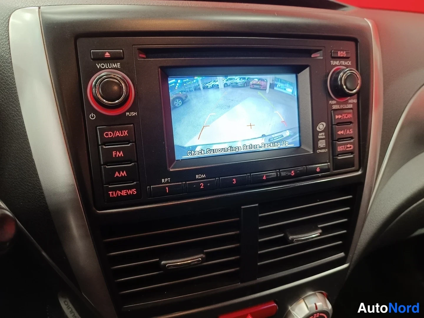Muu: Kojelaudan radiopaneeli lähikuvassa: näytössä peruutuskamera ja teksti 'Check Surroundings Before Backing Up', painikkeet CD/AUX, FM, AM, RPT, RDM ja numerot 1–
