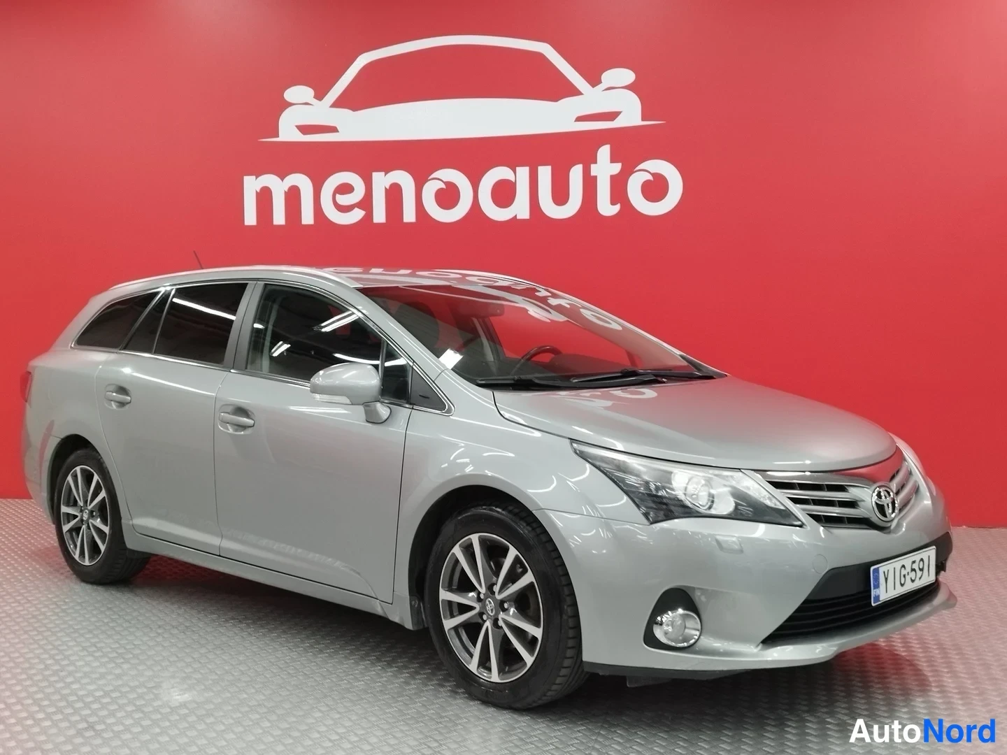Etuviisto, nokka oikealle: Hopeanharmaa Toyota Avensis -farmari etukulmassa, keulagrilla ja etupyörät näkyvissä punaisella taustalla.