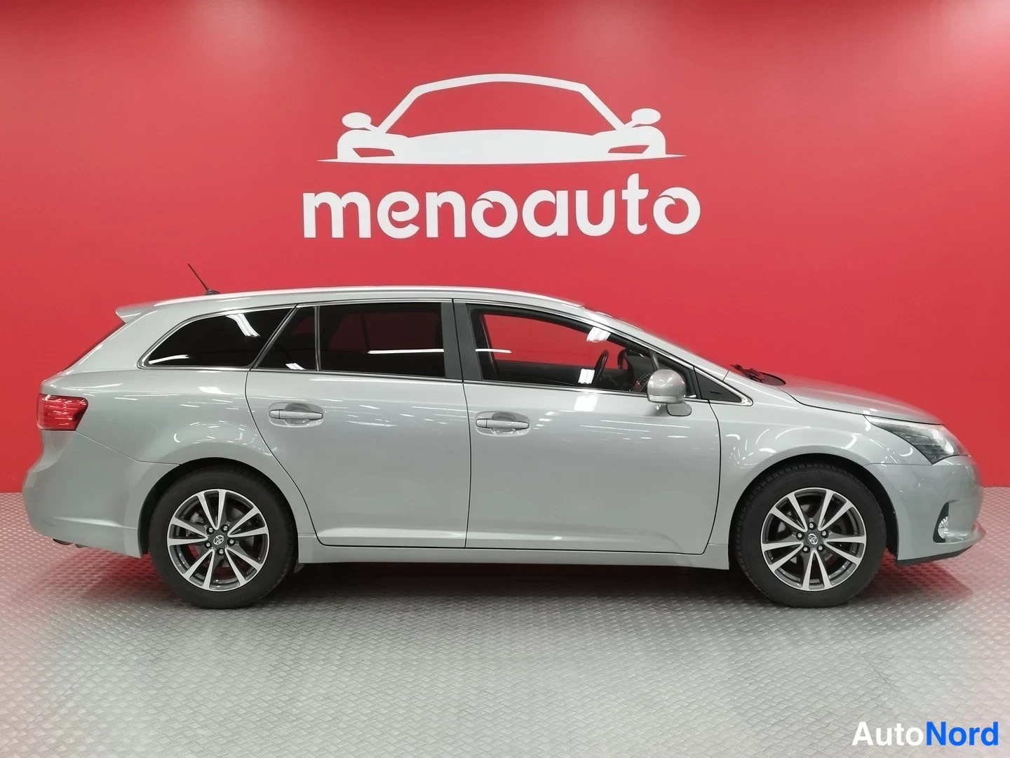 Sivulta: Hopeanharmaa Toyota Avensis -farmari sivukulmassa koko autotäydelään punaisen seinän edessä.