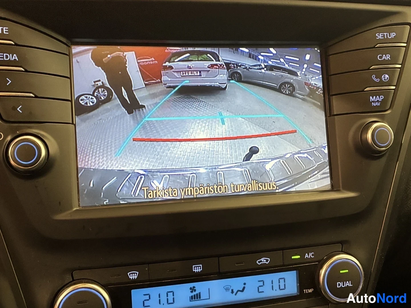 Yksityiskohta: Kojelaudan infotainment-näyttö näyttää peruutuskameran kuvan ohjausviivoineen ja tekstillä 'Tarkista ympäristön turvallisuus'. Näytön alla on ilmastointipaneeli
