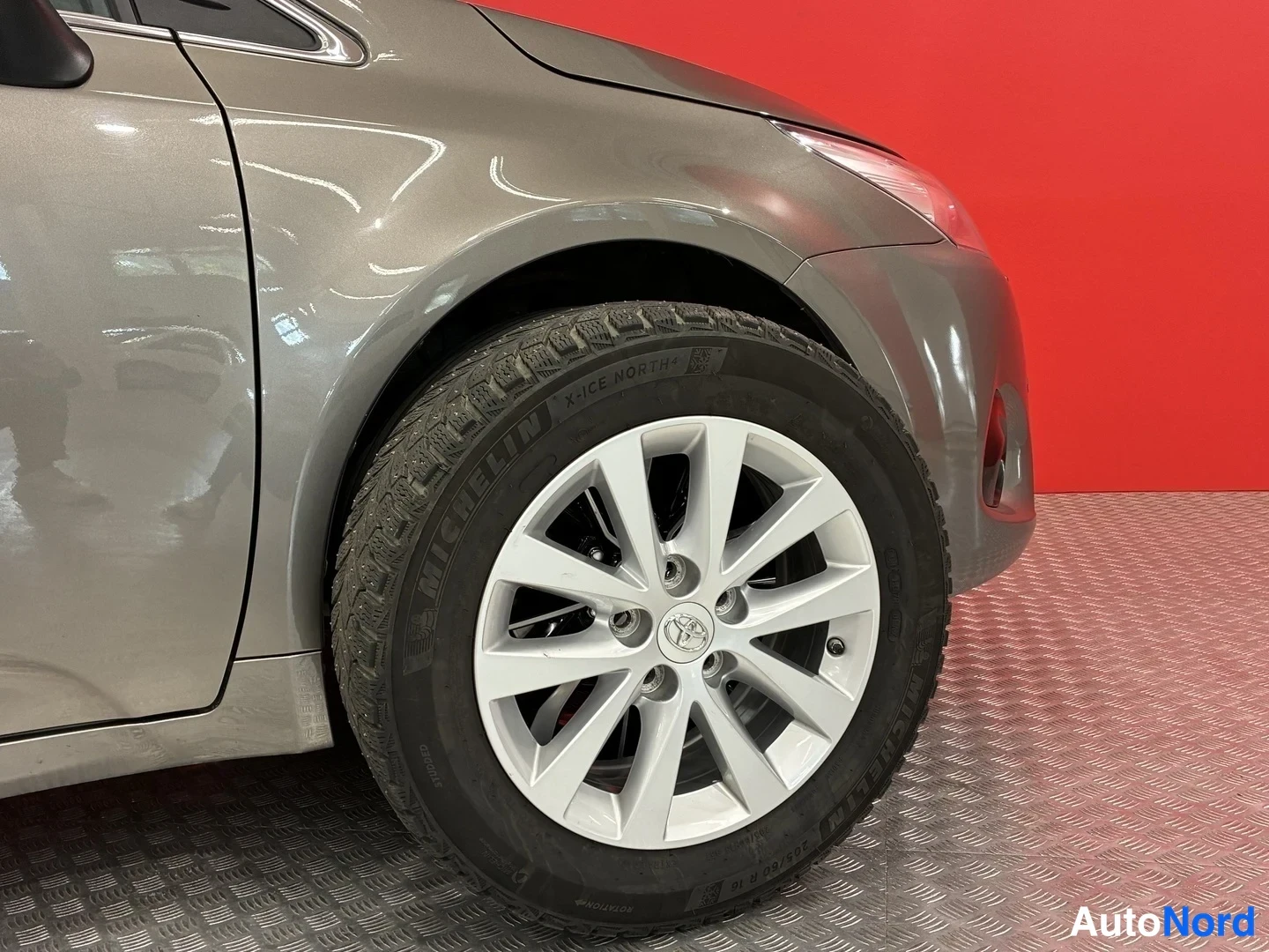 Etuviisto, nokka oikealle: Toyota Avensisin etukulma: etupyörä Michelin X-Ice North -nastarenkaalla ja hopeanharmaa kylkipaneeli punaista taustaa vasten.