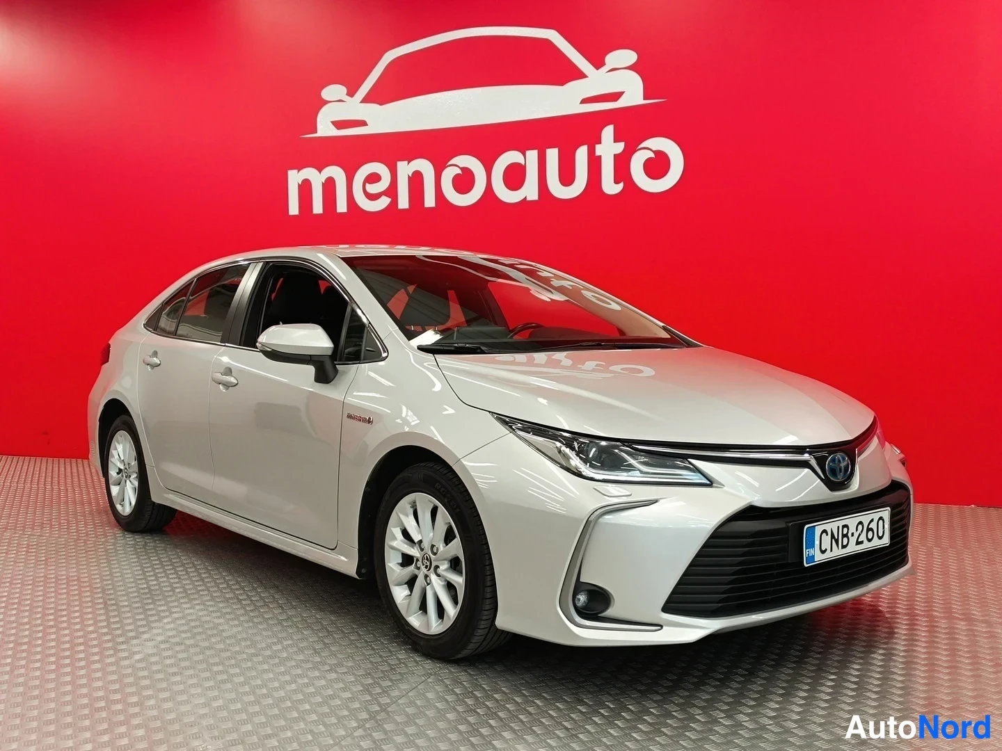 Etuviisto, nokka oikealle: Hopeanvärinen Toyota Corolla -sedan etukulmasta oikealta kuvattuna, rekisteritunnus, punainen menoauto-taustaseinä.