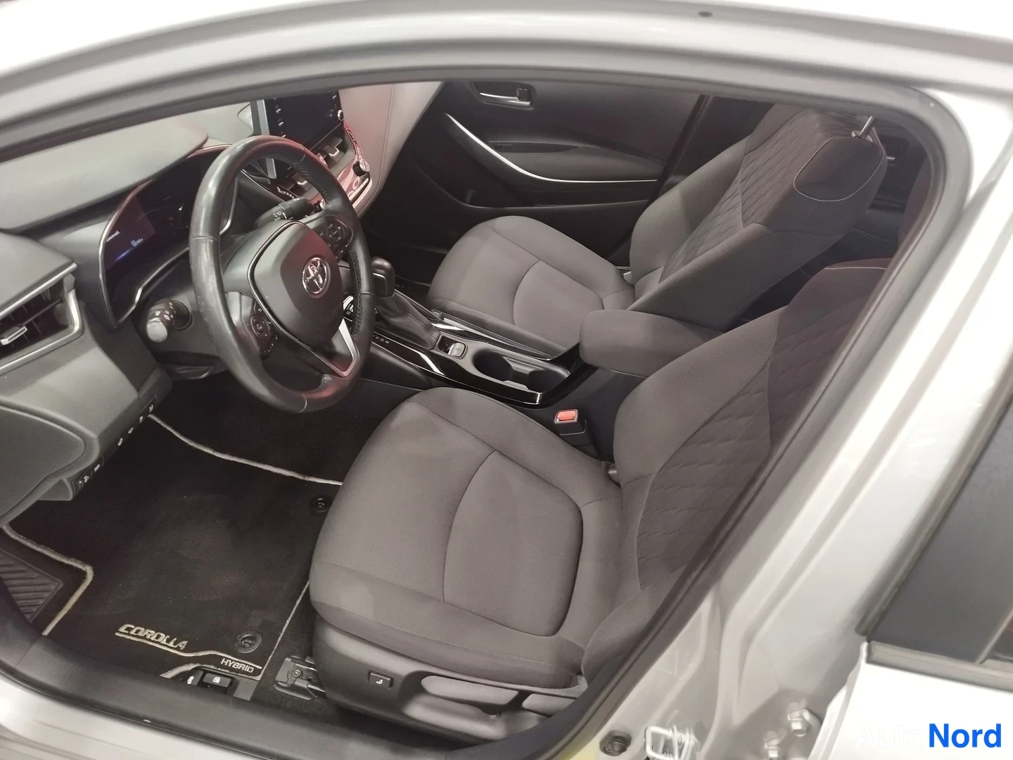Muu: Kuljettajan puolen ovi auki: Toyota-ohjauspyörä, automaattivaihteen valitsin ja tummat kangasistuimet, matolla COROLLA HYBRID -teksti.