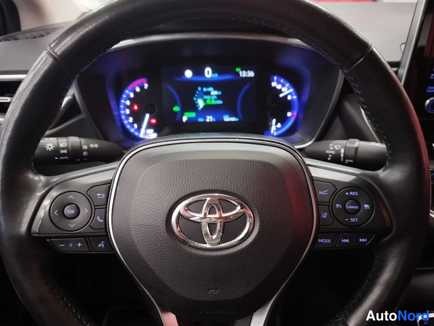 Edestä: Toyota-merkin omaava ohjauspyörä ja kojelaudan mittaristo, joissa näkyy hybridijärjestelmän valmiustila ja vakionopeudensäätimen painikkeet.