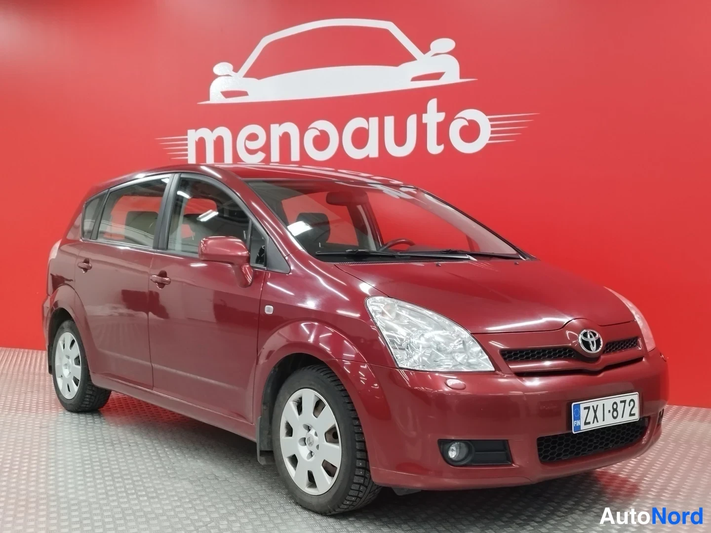 Etuviisto, nokka oikealle: Punainen Toyota Corolla Verso -tilavauto kuvattu etukulmasta, kylki ja keula näkyvissä, rekisterikilpi, punaisella taustalla.