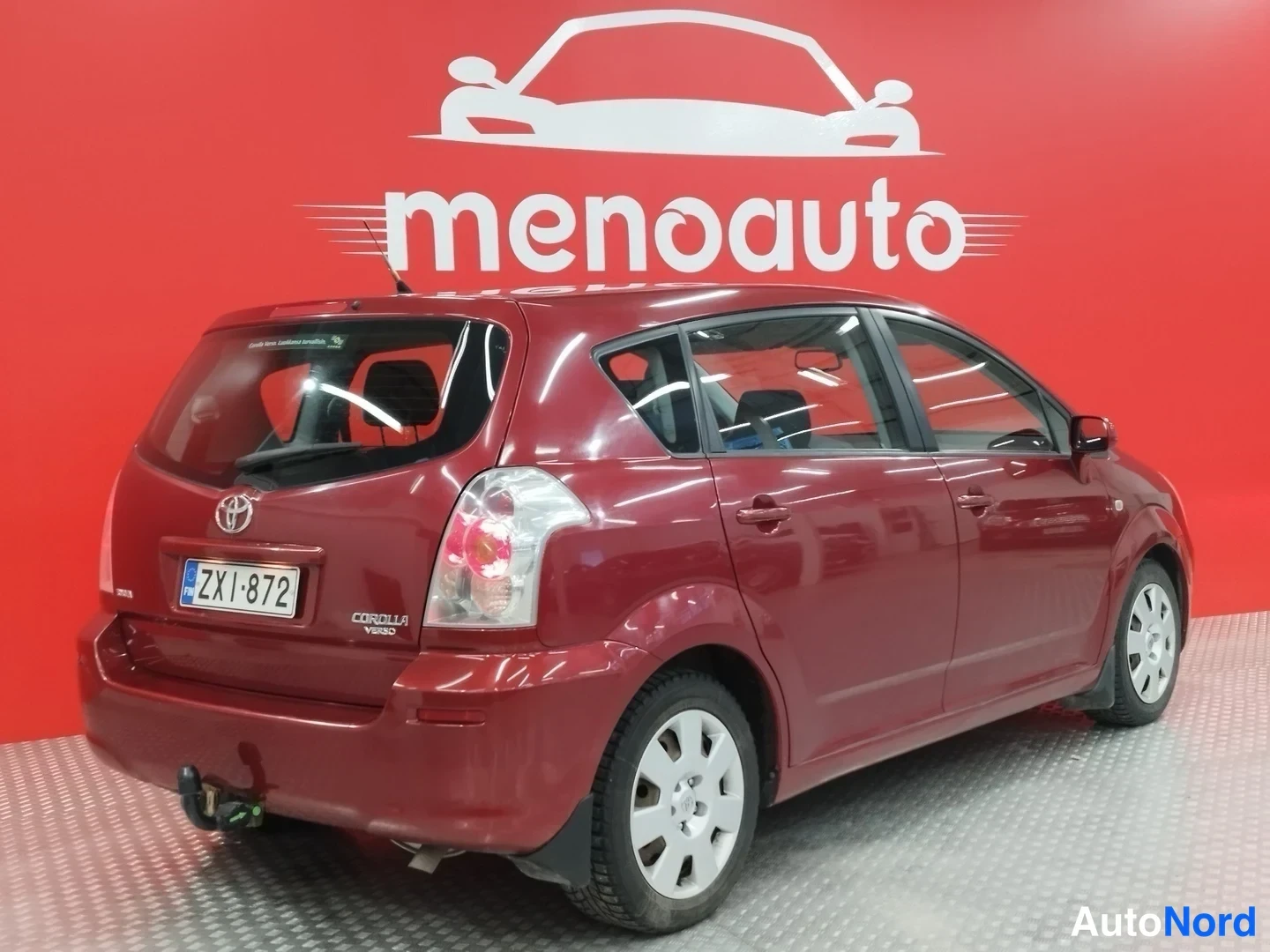 Takaviisto: Punaisen Toyota Corolla Verso -farmarin takakulma kuvattuna sisätiloissa punaisella taustalla; rekisterikilpi ja vetokoukku näkyvät.