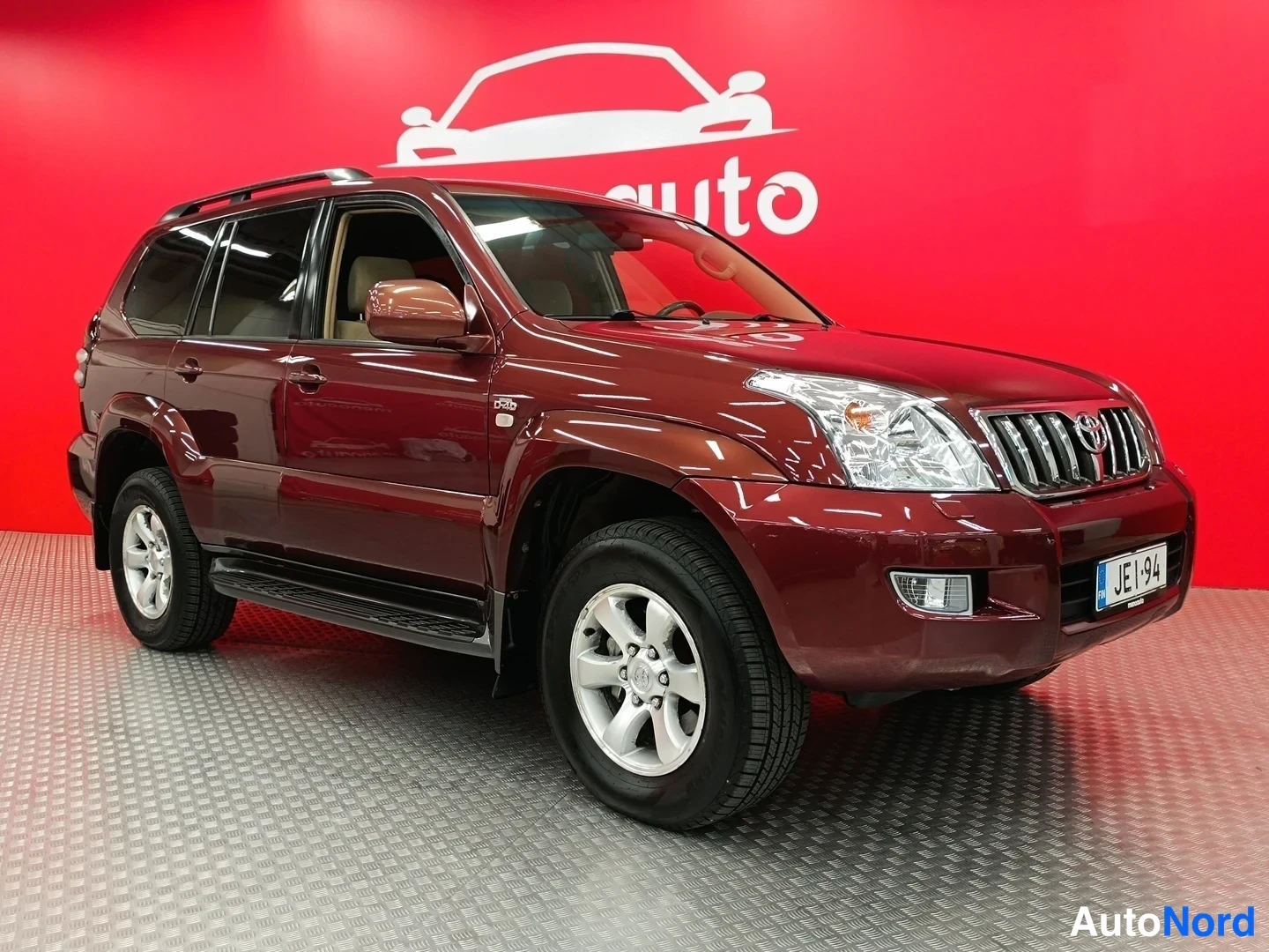 Etuviisto, nokka oikealle: Tummanpunaisen Toyota Land Cruiserin etukulma oikealta, kylki ja etumoitto näkyvät punaisella taustalla.