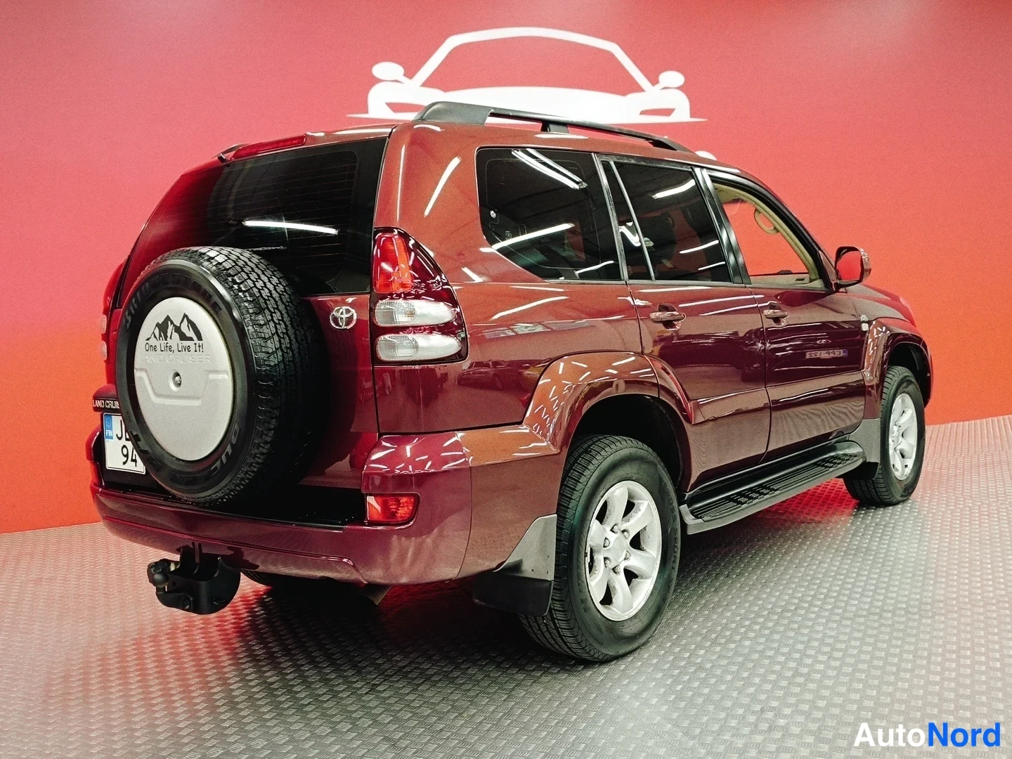 Takaviisto: Tummanpunaisen Toyota Land Cruiserin takakulma vasemmalta, varapyörä takaluukulla ja vetokoukku näkyvät.