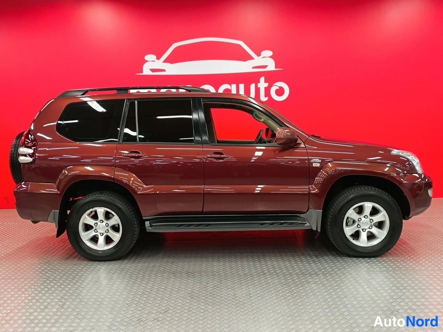 Etuviisto, nokka oikealle: Tummanpunaisen Toyota Land Cruiserin sivuprofiili oikealta puolelta, katolla kromikatokiskot ja sivuilla astinlaudat.
