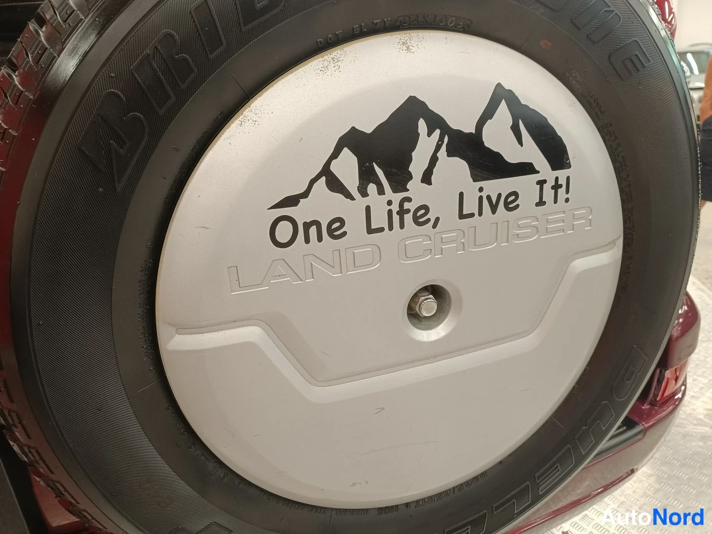 Yksityiskohta: Sivuteltu varapyörä auton perässä. Varapyörän kannessa lukee ONE LIFE, LIVE IT! ja sen alla LAND CRUISER -teksti, vuoristoraidoilla. Renaan kyljessä BRIDGESTONE