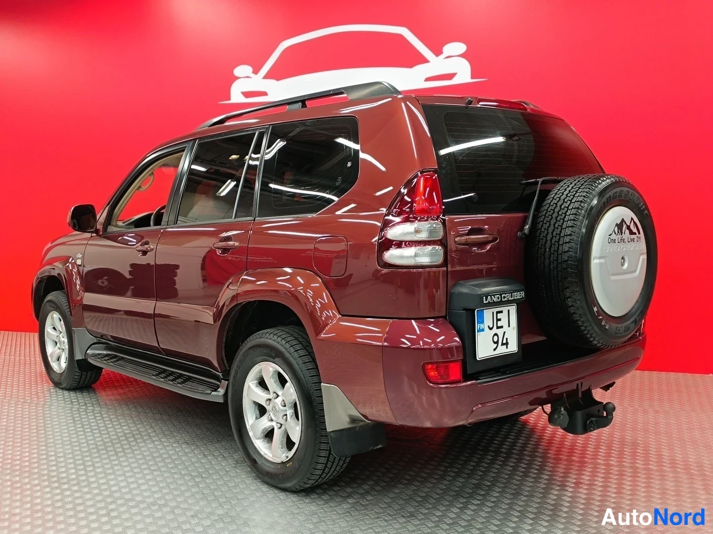 Takaviisto: Tummanpunaisen Toyota Land Cruiserin takaosa kolmannesnäkymästä, varatyökalu kannella, rekisteritunnus ja punainen tausta.