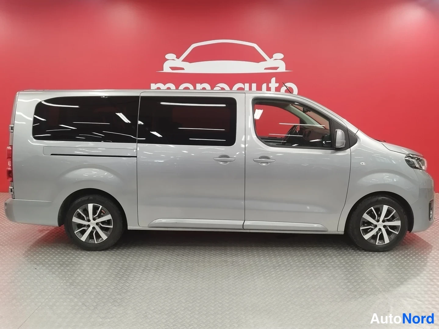 Etuviisto, nokka oikealle: Hopeaharmaa Toyota Proace Verso -pakettiauto sivulta oikealta, tummat takaikkunat ja kevytmetallivanteet näkyvät sisätiloissa punaisella taustalla.