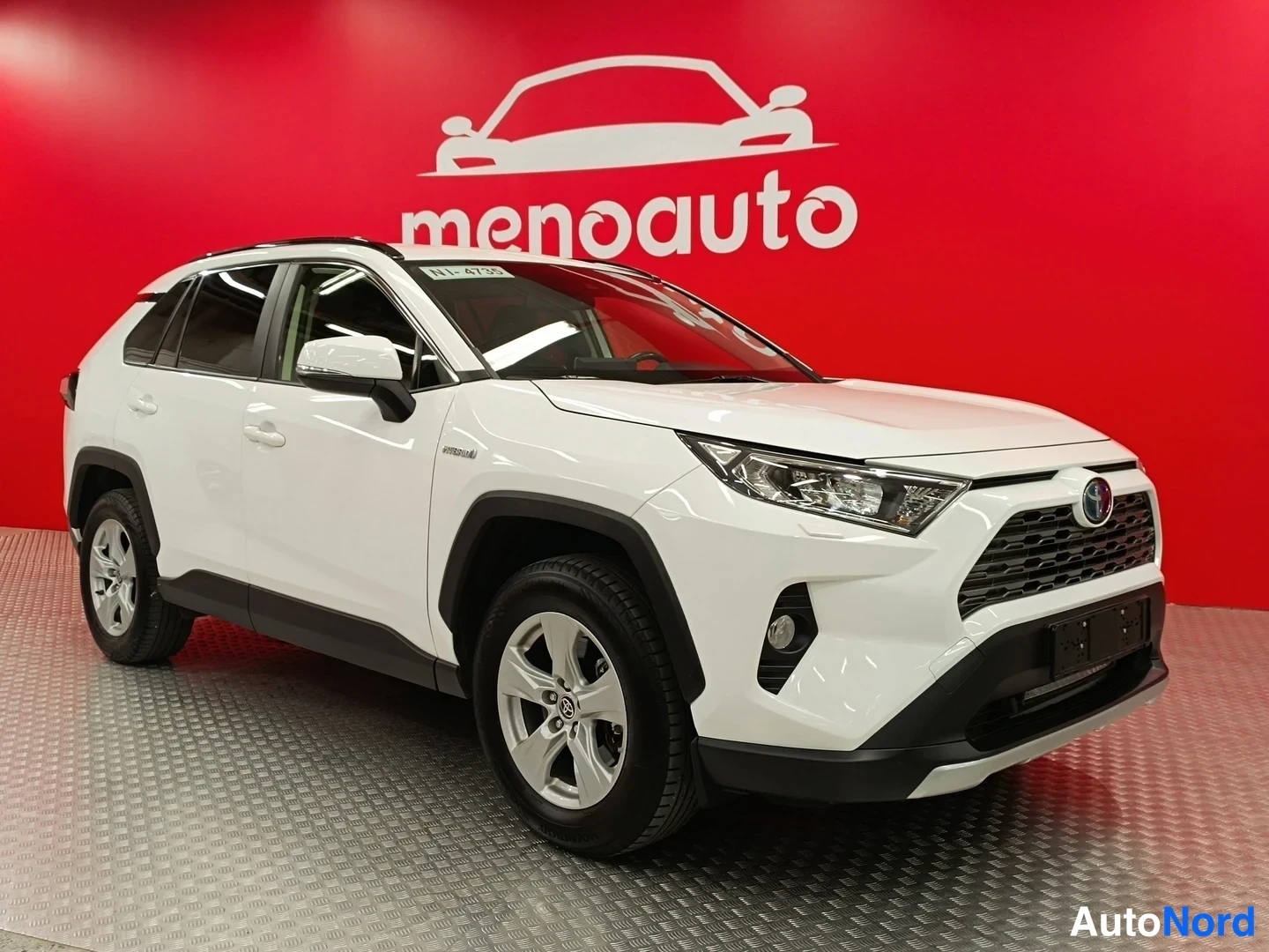 Etuviisto, nokka oikealle: Valkoinen Toyota RAV4 kulmassa edestä oikealta, etupanssi ja ajovalot näkyvissä punaisella menoauto-taustalla.