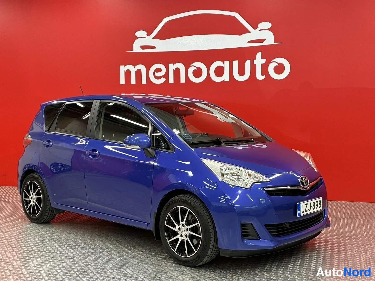 Etuviisto, nokka oikealle: Sininen Toyota Verso-S etkolta oikealta kuvattuna, rekisterikilpi, taustalla punainen menoauto-tunnaus.