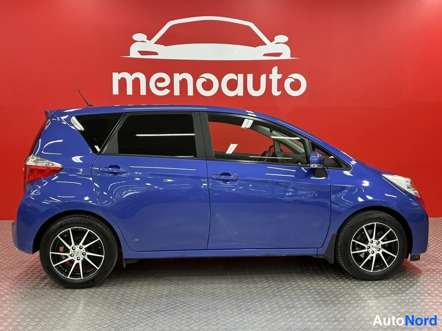 Sivulta: Sininen Toyota Verso-S sivulta vasemmalta kuvattuna punaisella menoauto-taustalla.