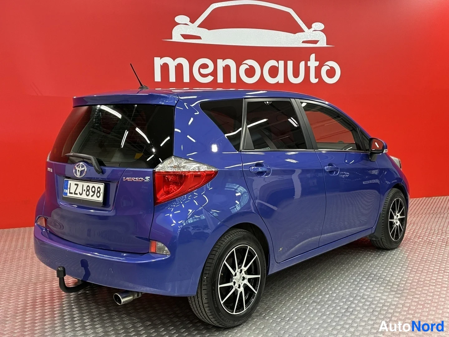 Takaviisto: Sininen Toyota Verso-S takakulmasta kuvattuna, rekisterikilpi ja lisävalo, taustalla menoauto-tunnaus.