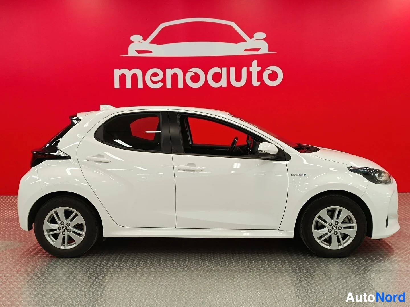 Sivulta: Valkoinen Toyota Yaris viistoperä sivulta vasemmalta kuvattuna, punainen menoauto-tausta.