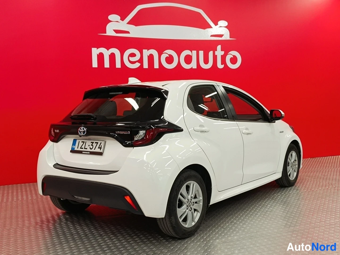 Takaviisto: Valkoinen Toyota Yaris viistoperä kuvattu kolmannelta osalta takaa vasemmalta, HYBRID-teksti takaluukussa, rekisteritunnus, punainen menoauto-tausta.