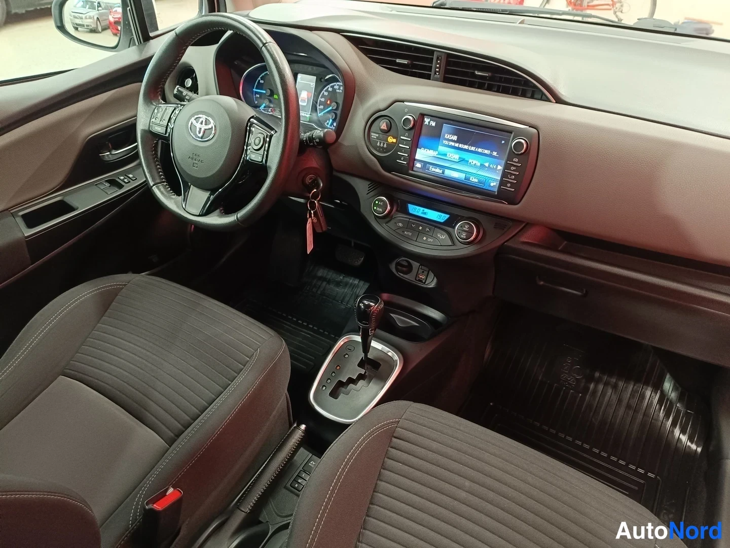 Muu: Kuljettajan näkökulma ohjaamosta:Toyota-ohjauspyörä,järjestelmänäyttö_RADIO-näyttö päällä,automaattivalitsin ja kaksi tummaa kangasistuinta.