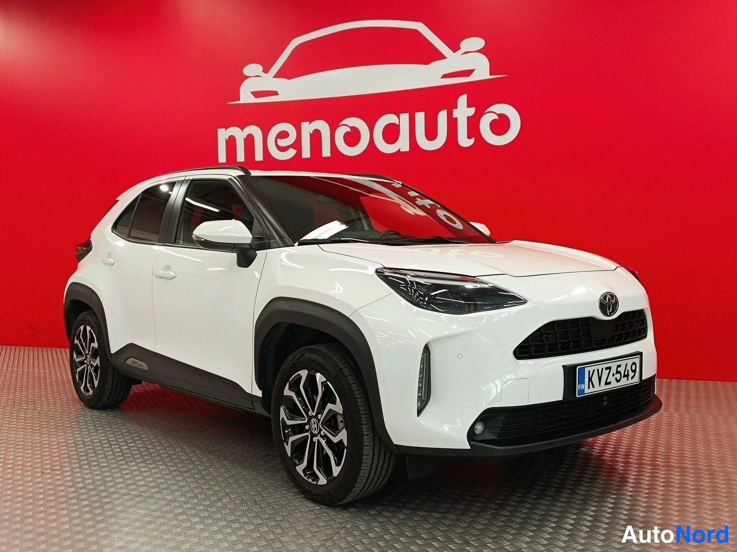 Etuviisto, nokka oikealle: Valkoinen Toyota Yaris Cross -maastokohtea kulmassa etureuna oikealle kuvattuna punaisella taustalla, rekisteritunnus.