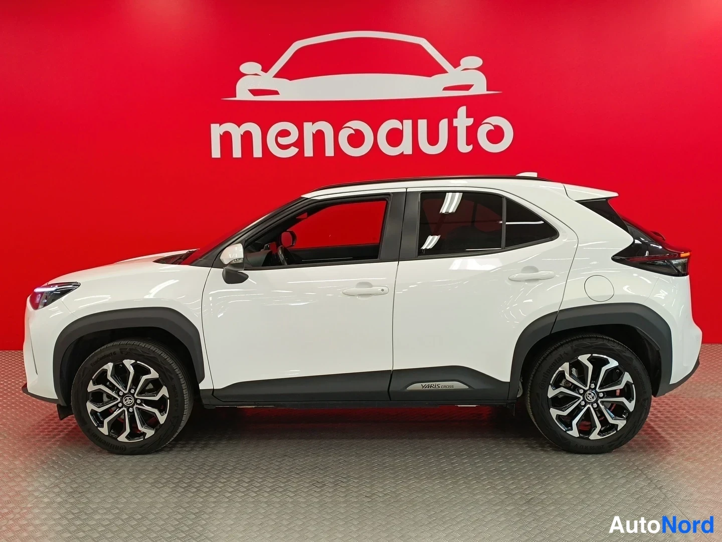 Etuviisto, nokka vasemmalle: Valkoinen Toyota Yaris Cross -tyyppinen maastocompakti sivulta-etukulmasta, nokka vasemmalla, punainen menoauto-tausta.
