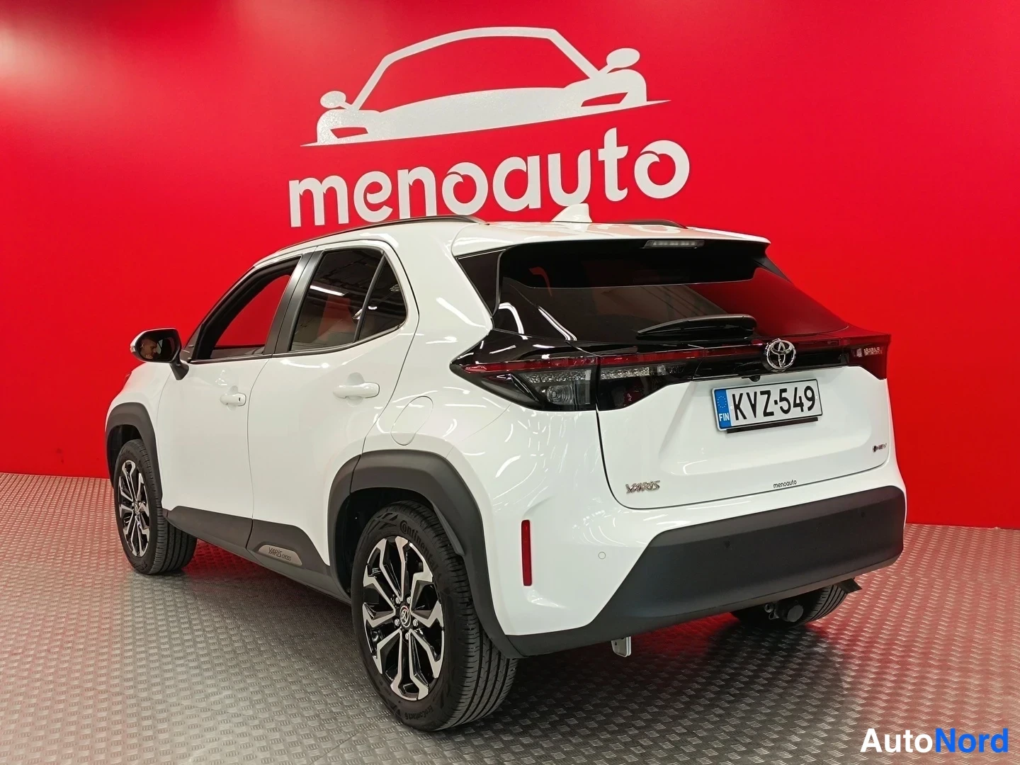 Takaviisto: Valkoisen Toyota Yaris Crossin takakulma rekisterikilvellä, punainen menoauto-tausta.