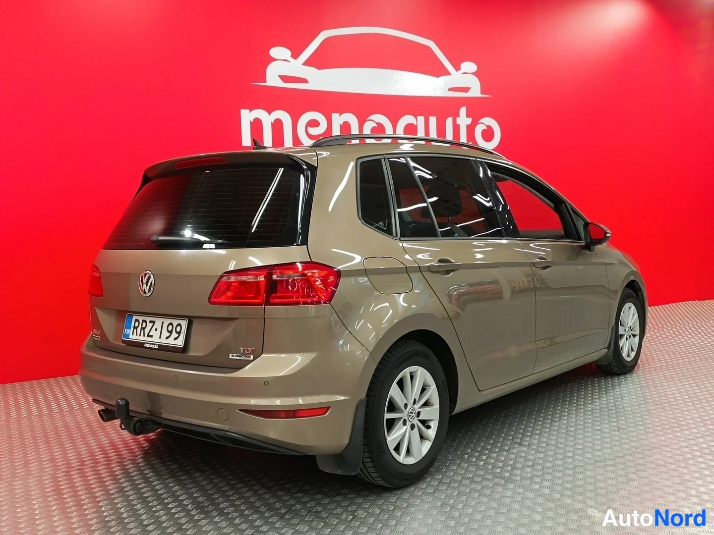 Takaviisto: Volkswagen Golf Sportsvan -farmarin takakulmakuva vasemmalta, punaiset takavalot, rekisteritunnus, vaaleanruskea koripelti, punainen menoauto-taustaseinä.