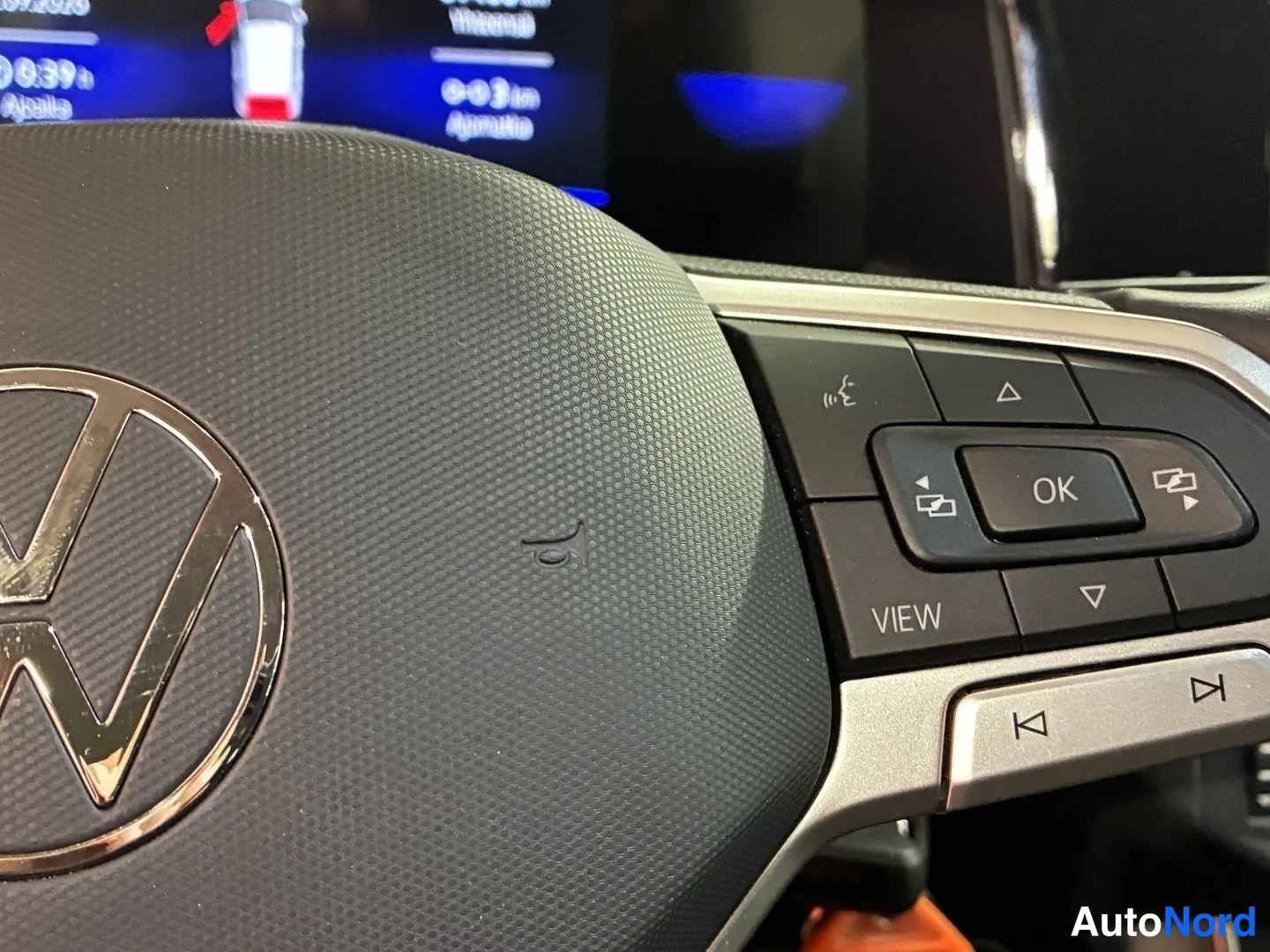 Yksityiskohta: Sisäkuva Volkswagenin ohjauspyörästä: keskellä kromattu VW-logo, oikealla pyörän monitoiminäppäimet (VIEW, OK, valikko- ja ääniohjausnäppäimet), taustalla ositt