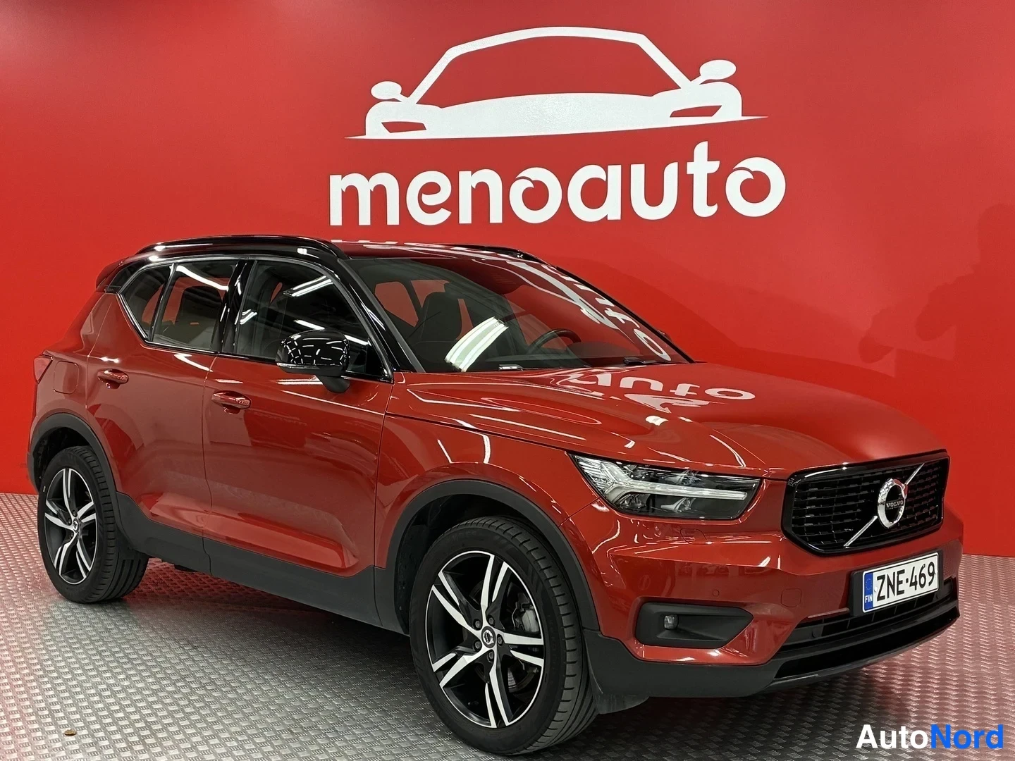 Etuviisto, nokka oikealle: Punainen Volvo XC40 -maastoauto etuviistosta oikealta kuvattuna, rekisteritunnus, musta katto ja tummat vanteet, valokuvattu sisätiloissa punaisella sein